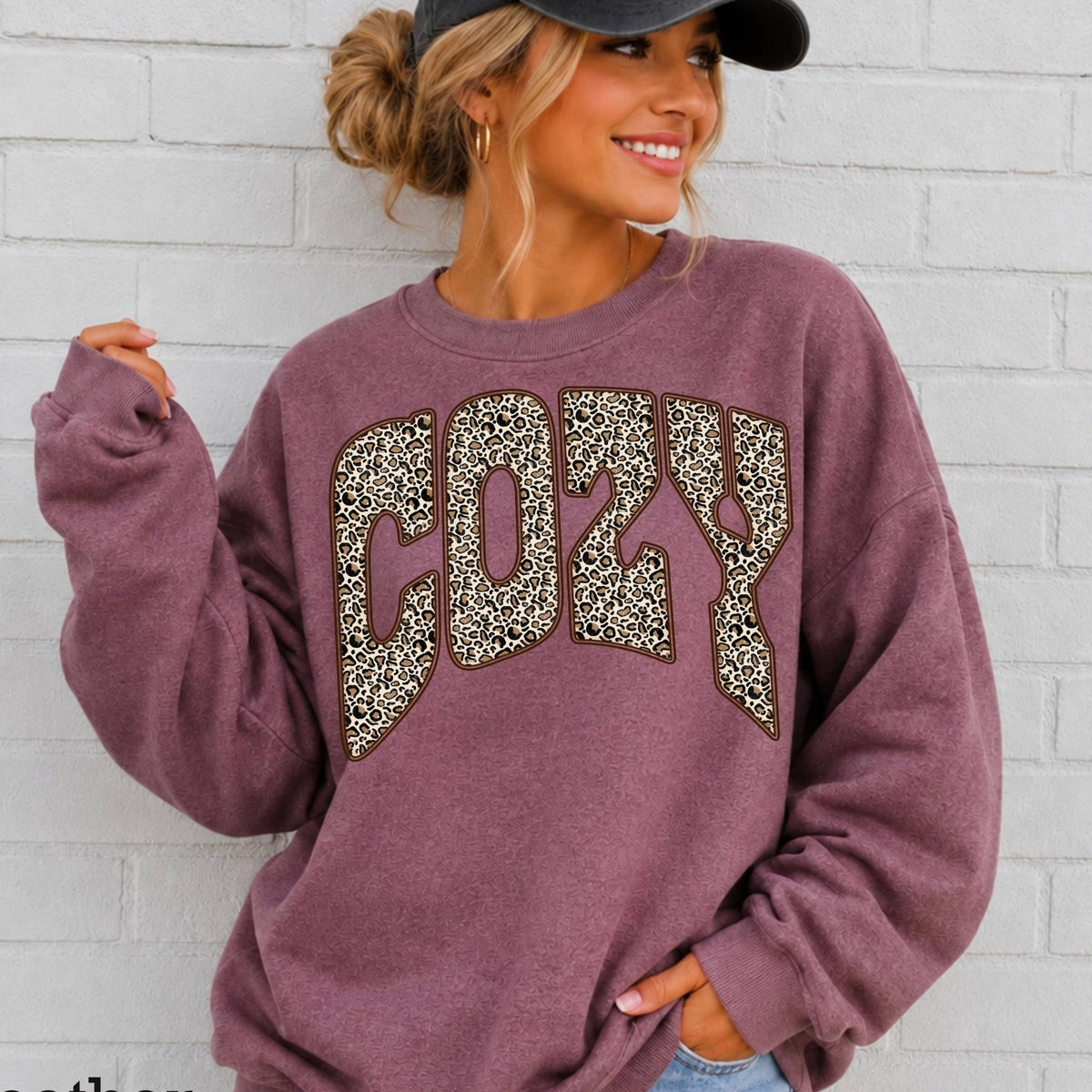 A woman wearing a heather maroon sweatshirt with a leopard-print 'COZY' graphic on the front, paired with a black baseball cap and light-wash jeans, standing against a white brick wall. Text in the bottom-left corner reads 'Heather Maroon'.
