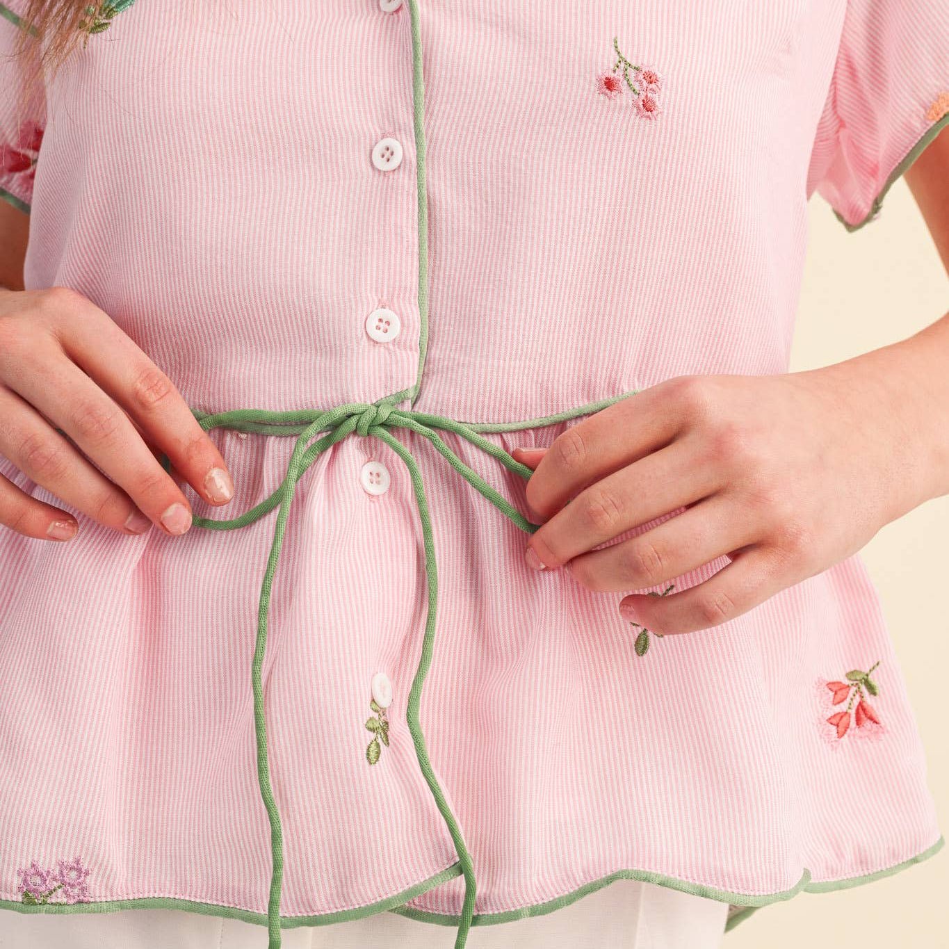 Close-up of a person wearing a pink outfit with floral embroidery and a green tie, on a light yellow background.