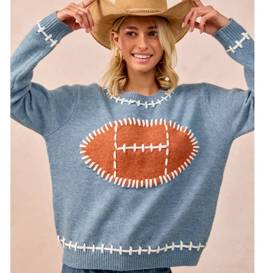 A woman wearing a light blue knit sweater with a large brown football applique on the chest and white stitched details, paired with a tan cowboy hat and blue jeans, against a plain beige studio background.
