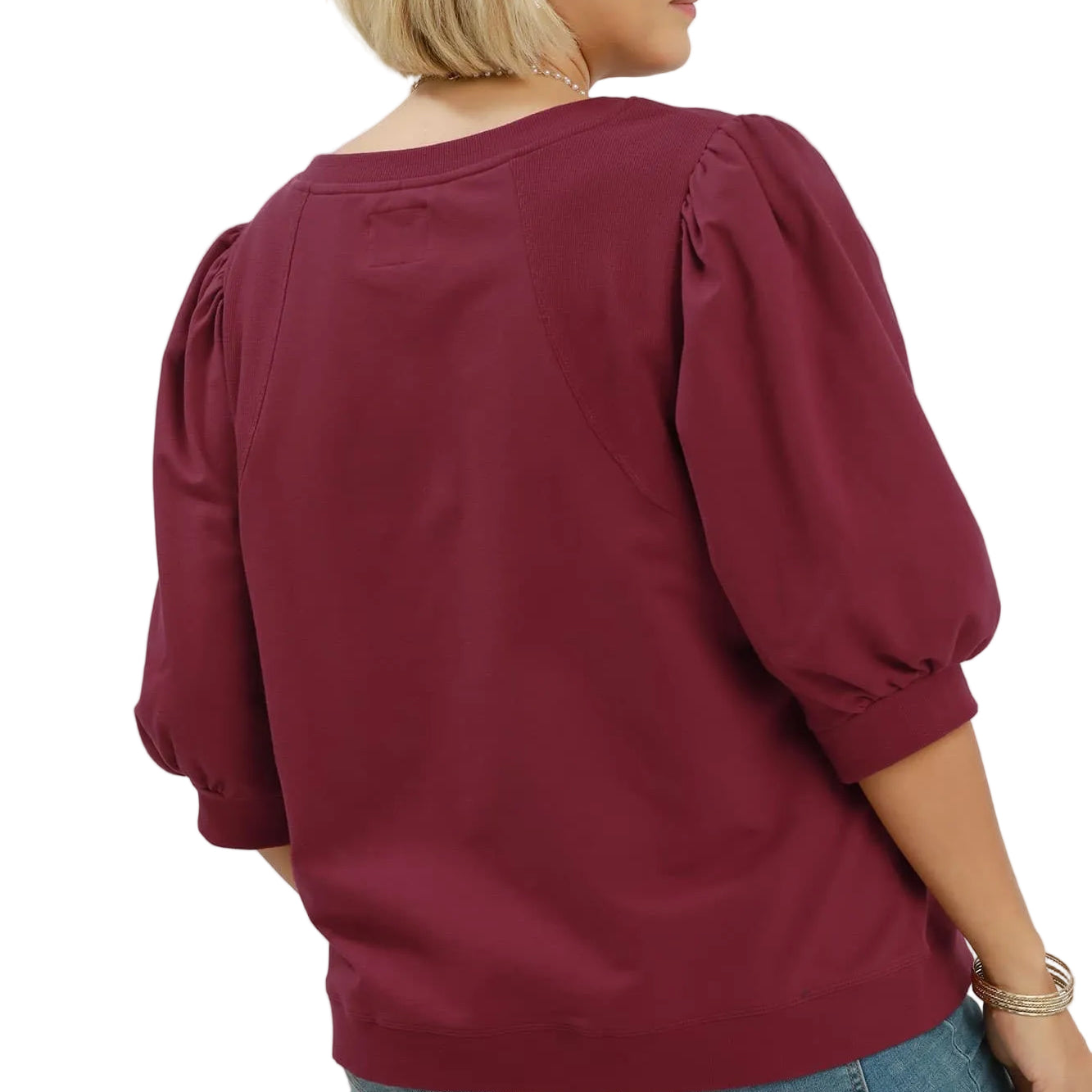 A woman shown from the back wearing a wine-colored puff-sleeve blouse and blue jeans against a light gray studio background. The text 'PLUSSIZE' appears in the top right, and 'WINE' appears in the bottom right.