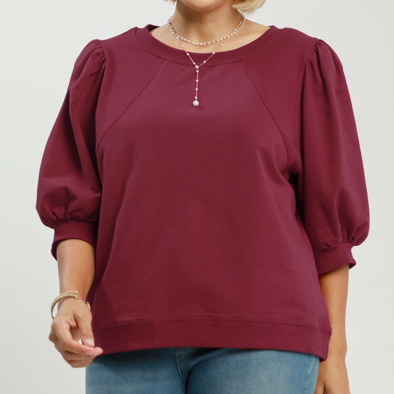 A woman modeling a wine-colored long-sleeve top with voluminous puff sleeves and a round neckline, paired with blue jeans. The image has a white background with 'PLUS SIZE' text in the top right and 'WINE' text in the bottom right.