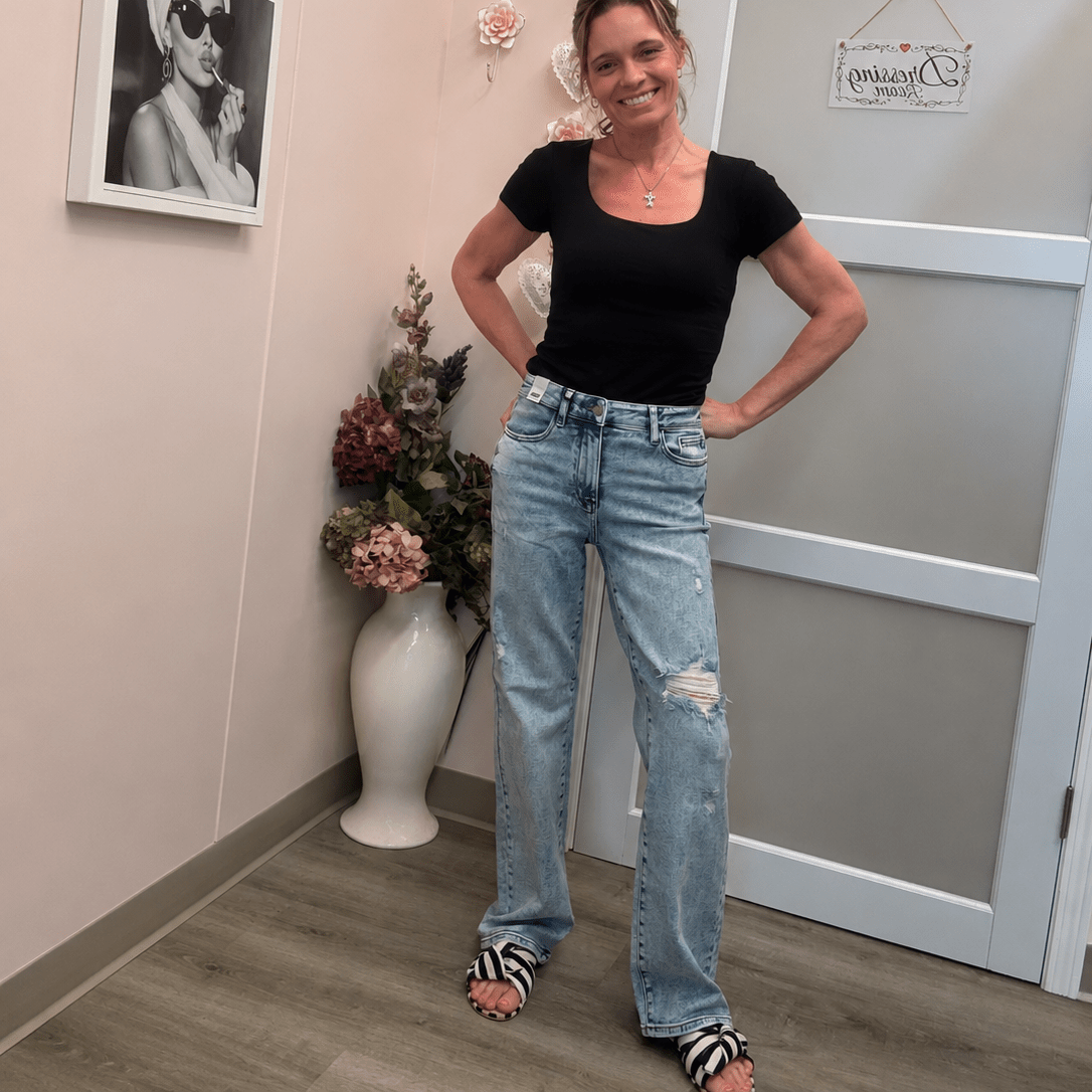 A woman posing in a black scoop-neck t-shirt and light-wash distressed jeans inside a room with pink walls, framed art, and a white door.