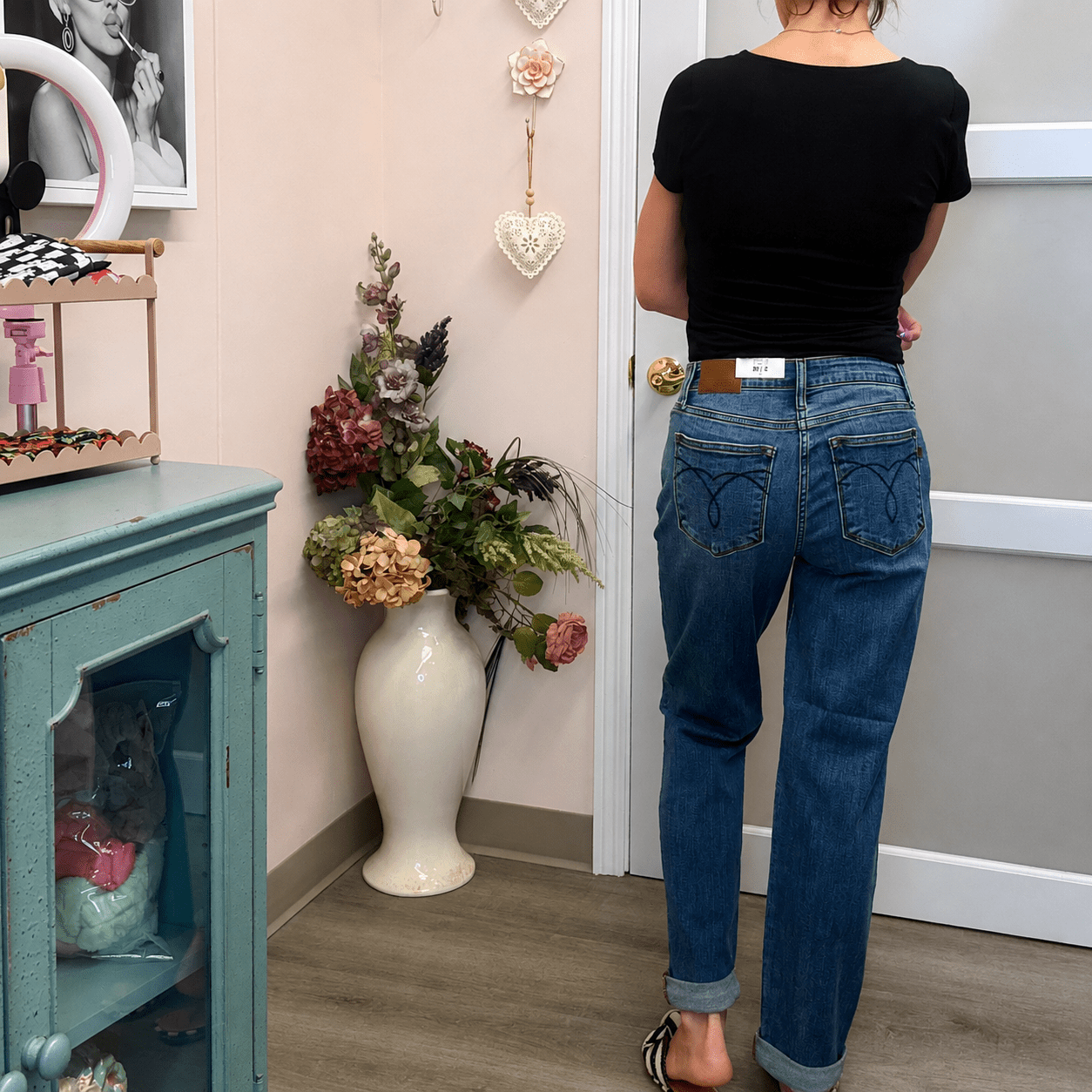 A person viewed from behind wearing blue jeans with decorative stitched back pockets and a black t-shirt, standing in a home interior next to a teal cabinet and a vase of flowers.
