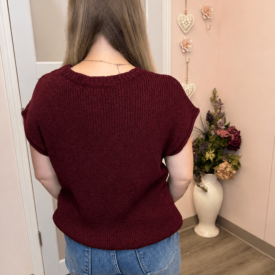 A woman viewed from behind wearing a short-sleeve ribbed knit sweater in burgundy and blue jeans, standing in a room with a pink wall, wooden floor, and a vase of flowers.