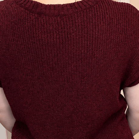 A woman viewed from behind wearing a short-sleeve ribbed knit sweater in burgundy and blue jeans, standing in a room with a pink wall, wooden floor, and a vase of flowers.