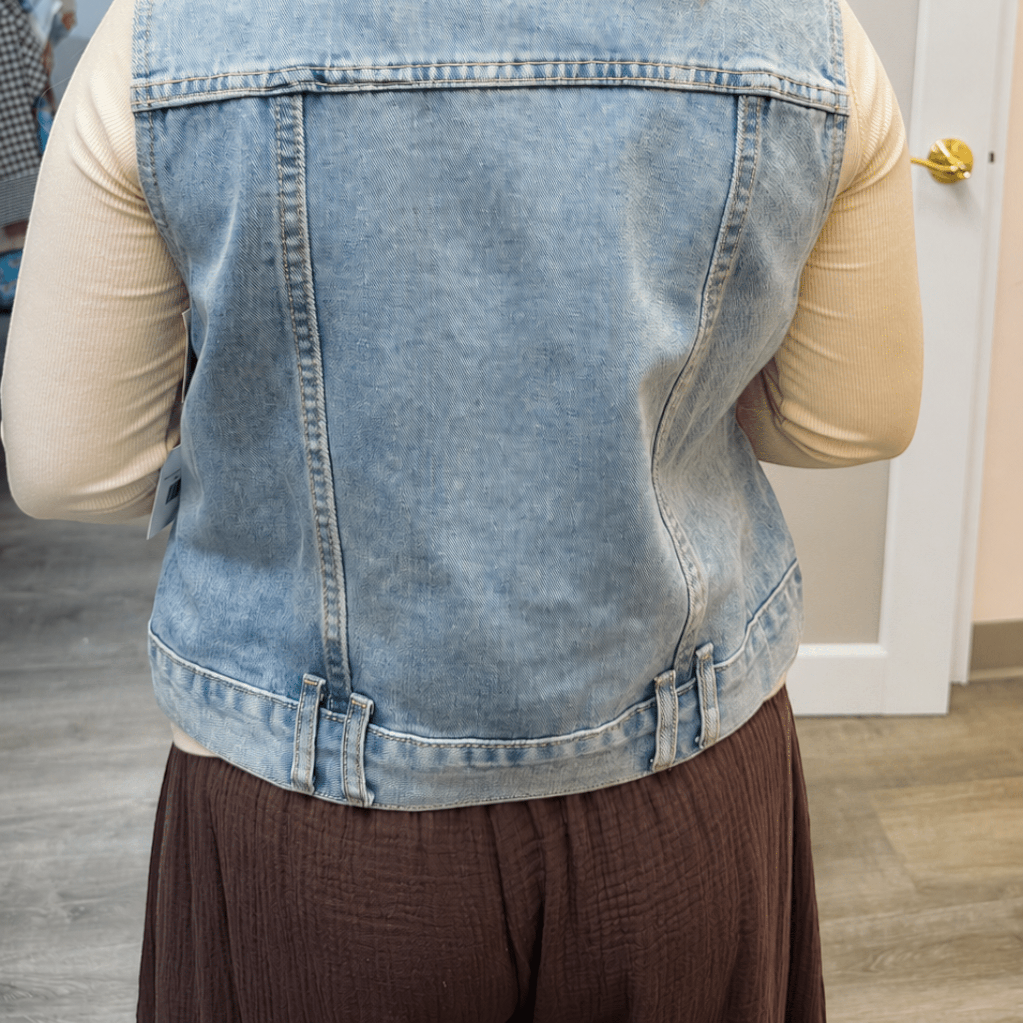 A person viewed from behind wearing a light blue denim vest over a beige long-sleeve top and dark brown pants, standing indoors near a white door.