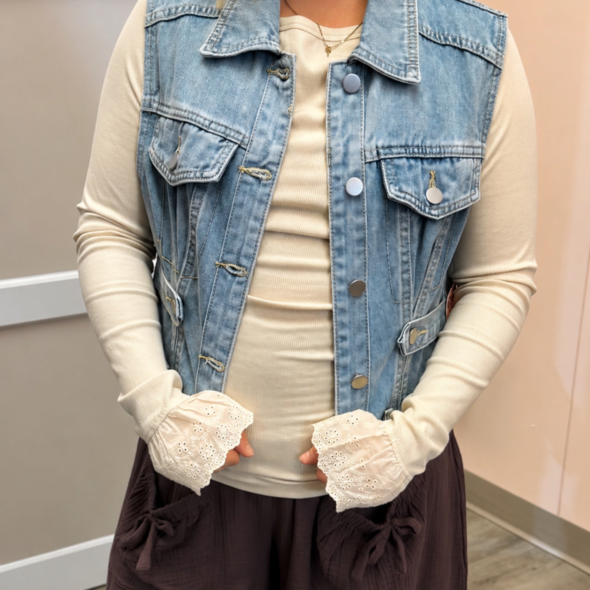 A person wearing a light blue denim vest over a cream long-sleeve top, with cream eyelet fingerless gloves and dark brown bottoms, standing indoors against a neutral wall.