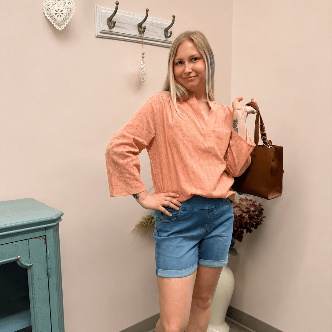 girl in peach top with light denim shorts is standing holding a brown purse.