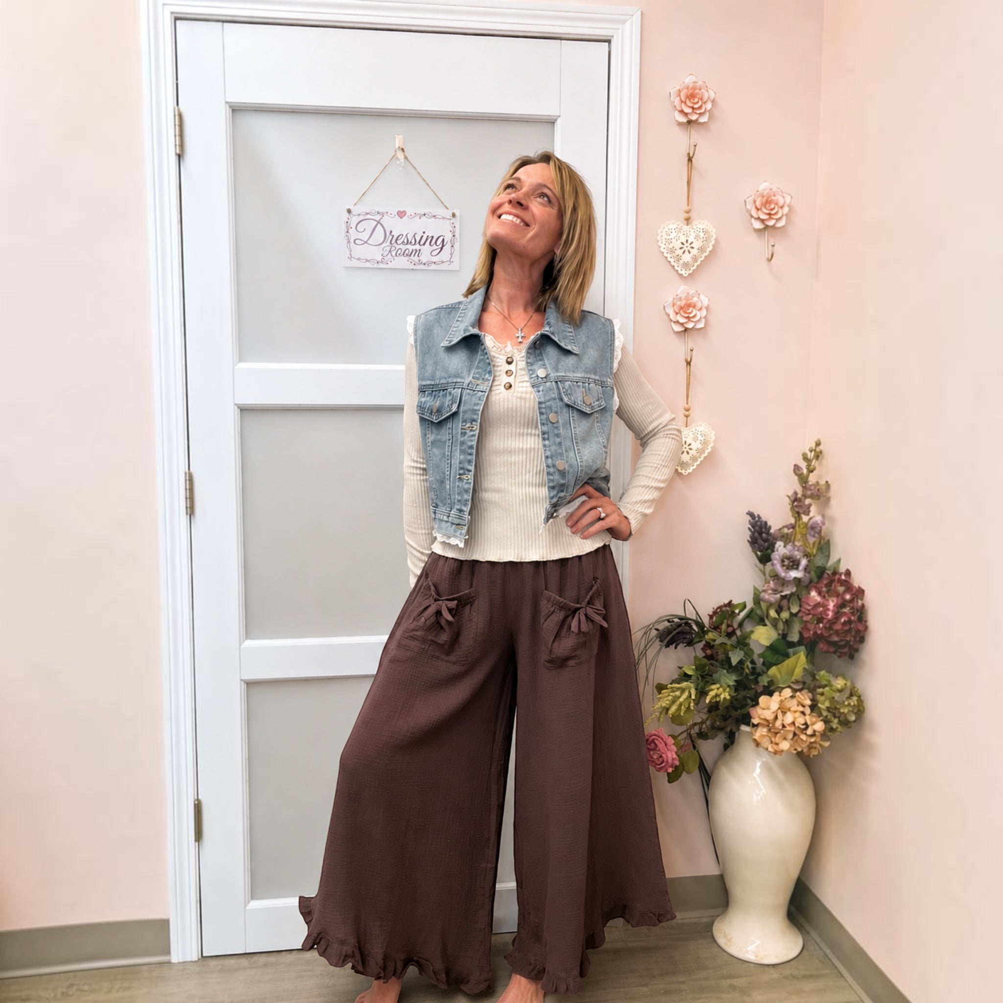 A woman modeling a casual outfit consisting of a light-wash denim vest over a cream ribbed top and brown wide-leg pants with bow details, standing in a room with a pink wall, white door, and a vase of flowers.
