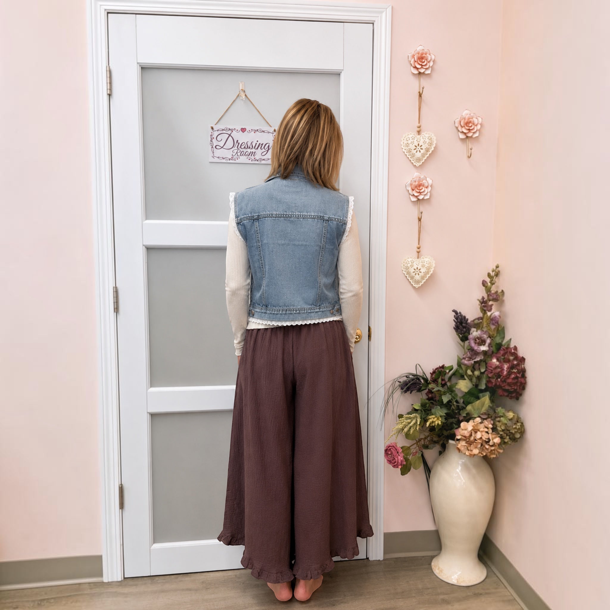 A woman stands with her back to the camera in front of a white door, wearing a light blue denim vest over a white long-sleeve top and a long brown skirt. The room has a pink wall decorated with hanging flowers and a vase of dried flowers.