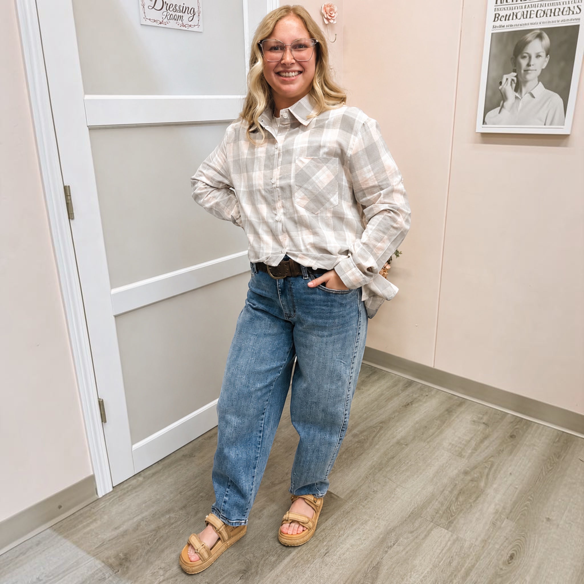 A woman stands indoors wearing a light beige and white plaid button-up shirt, blue jeans, and tan sandals. The setting appears to be a dressing room or fitting space with a door, framed wall art, and wood-look flooring.