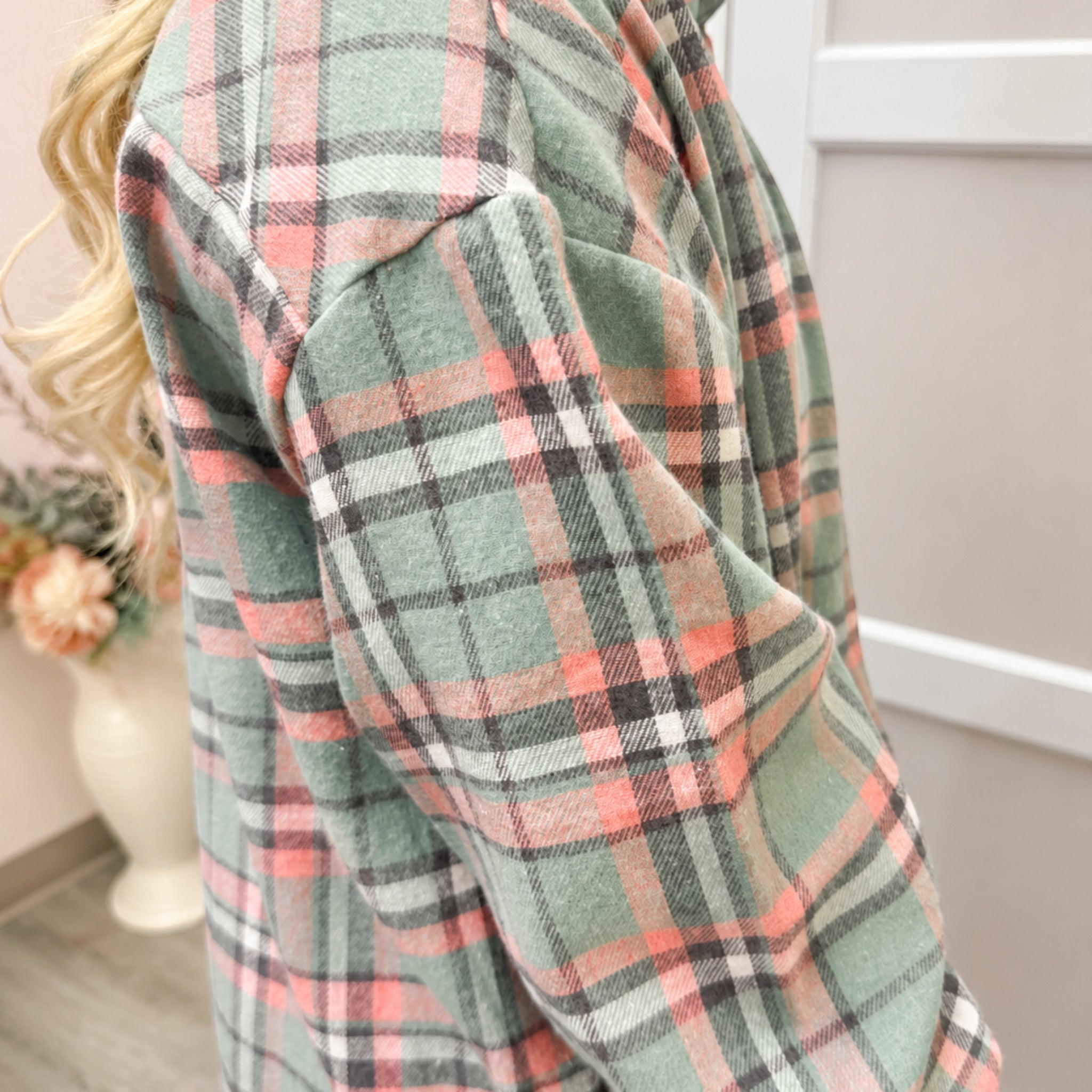 A close-up of a person wearing a long-sleeve plaid flannel shirt in sage green, pink, black, and white. The background shows a neutral indoor room with a vase of flowers.