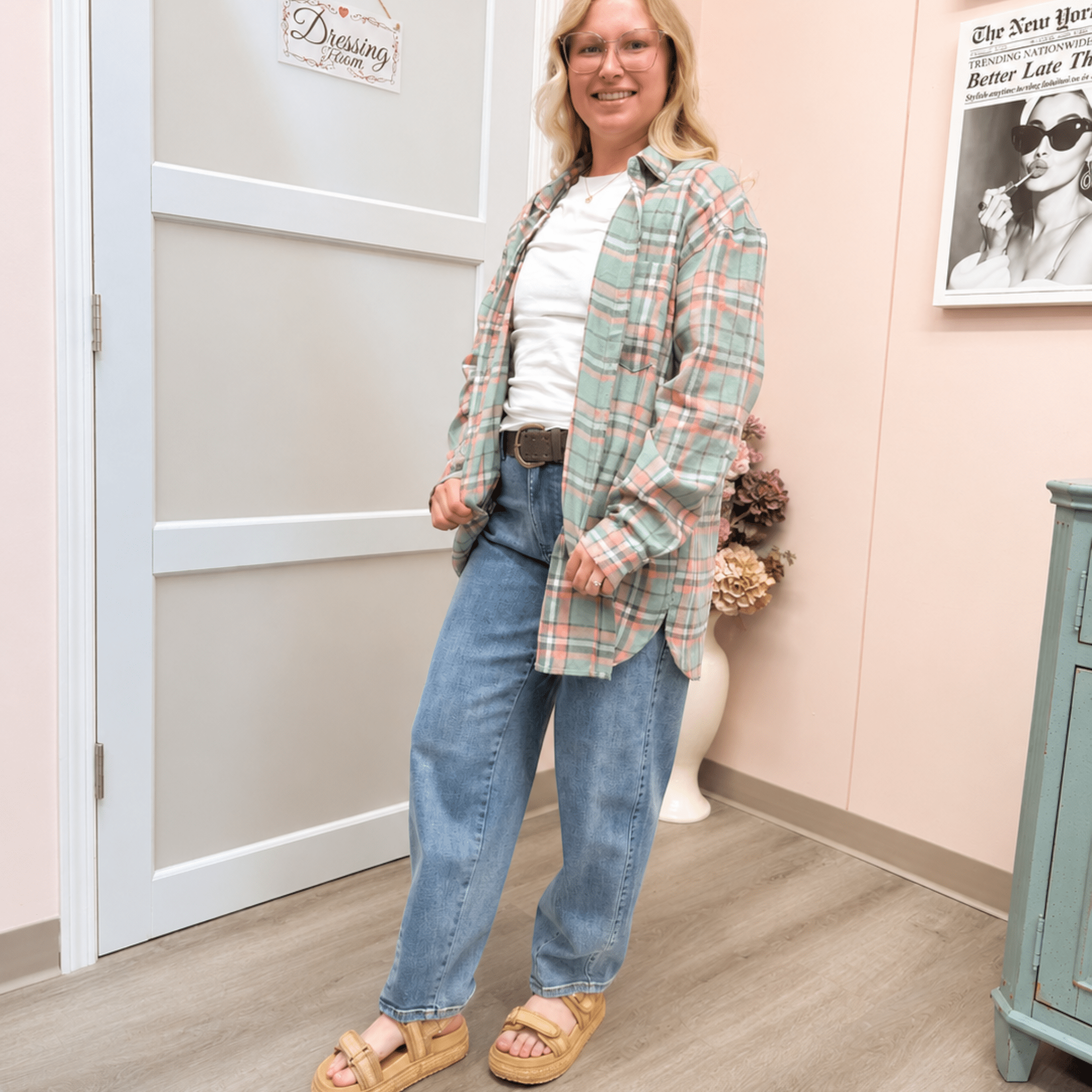 A woman modeling a light green and pink plaid button-up shirt over a white t-shirt, paired with blue jeans and tan platform sandals, standing in a room with a pink wall and a white door.