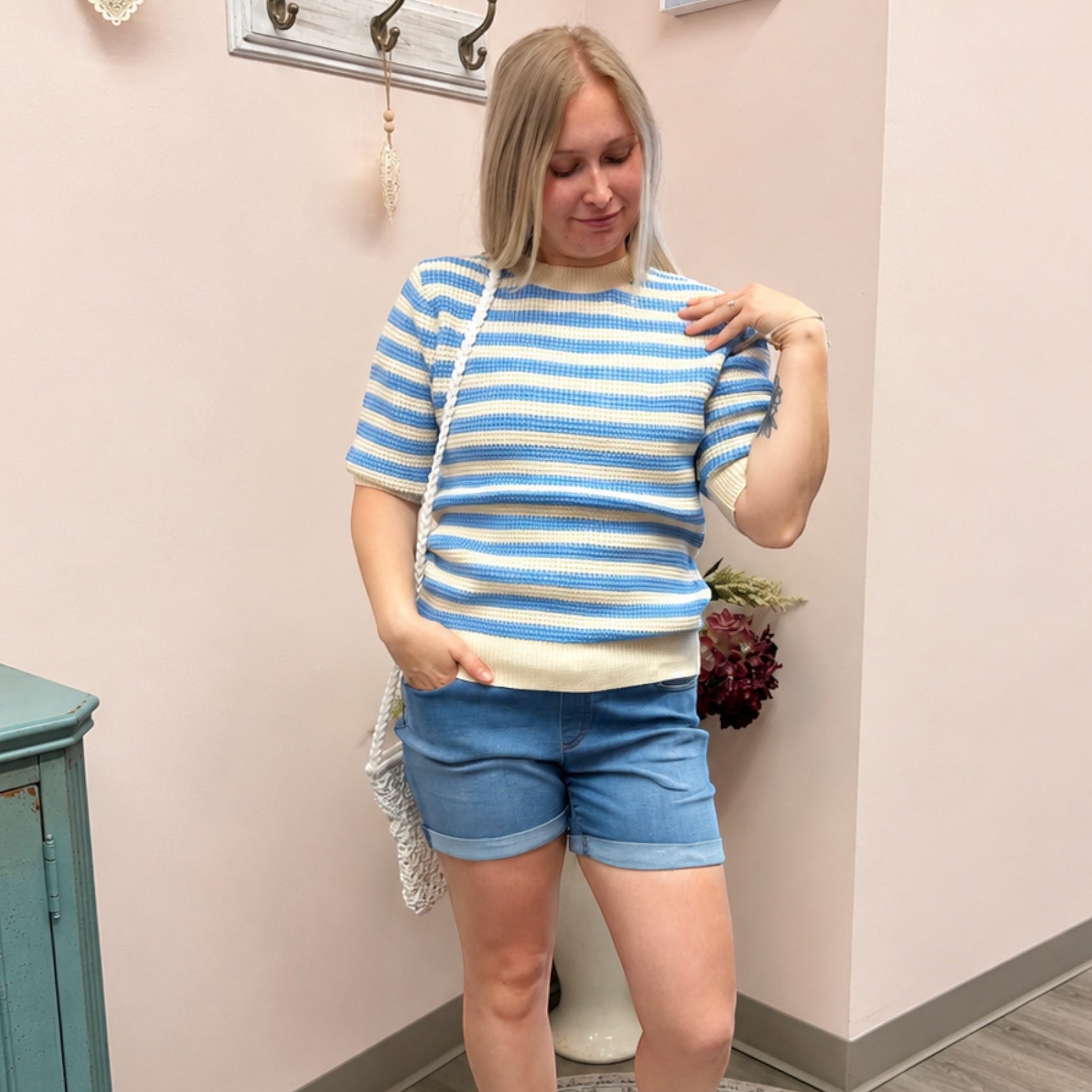 Woman wearing a blue and white striped shirt and shorts standing in a room with a light pink wall.