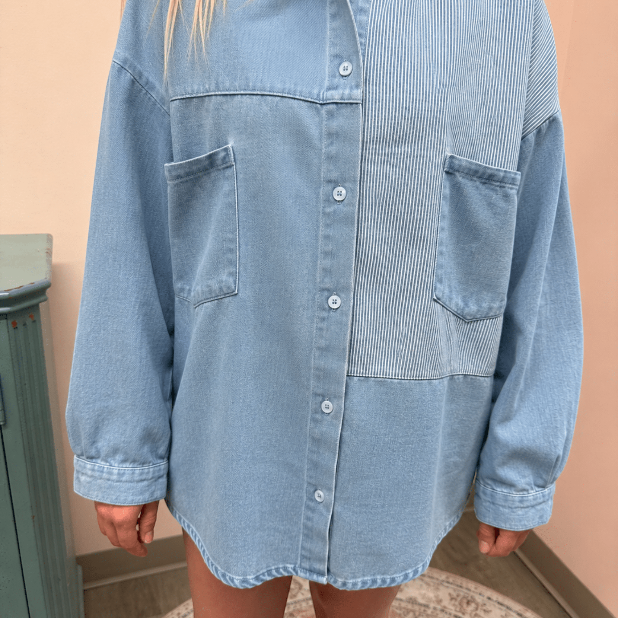 Person wearing a light blue denim shirt over a striped shirt against a beige wall.