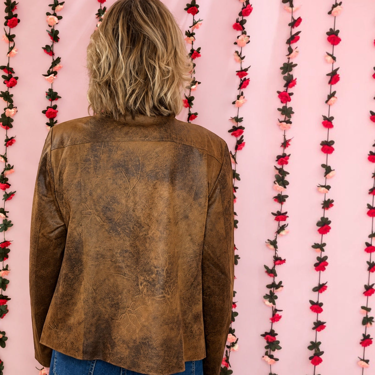 A person viewed from behind wearing a brown distressed leather jacket and blue jeans, standing against a pink backdrop decorated with vertical strands of red and pink artificial flowers.