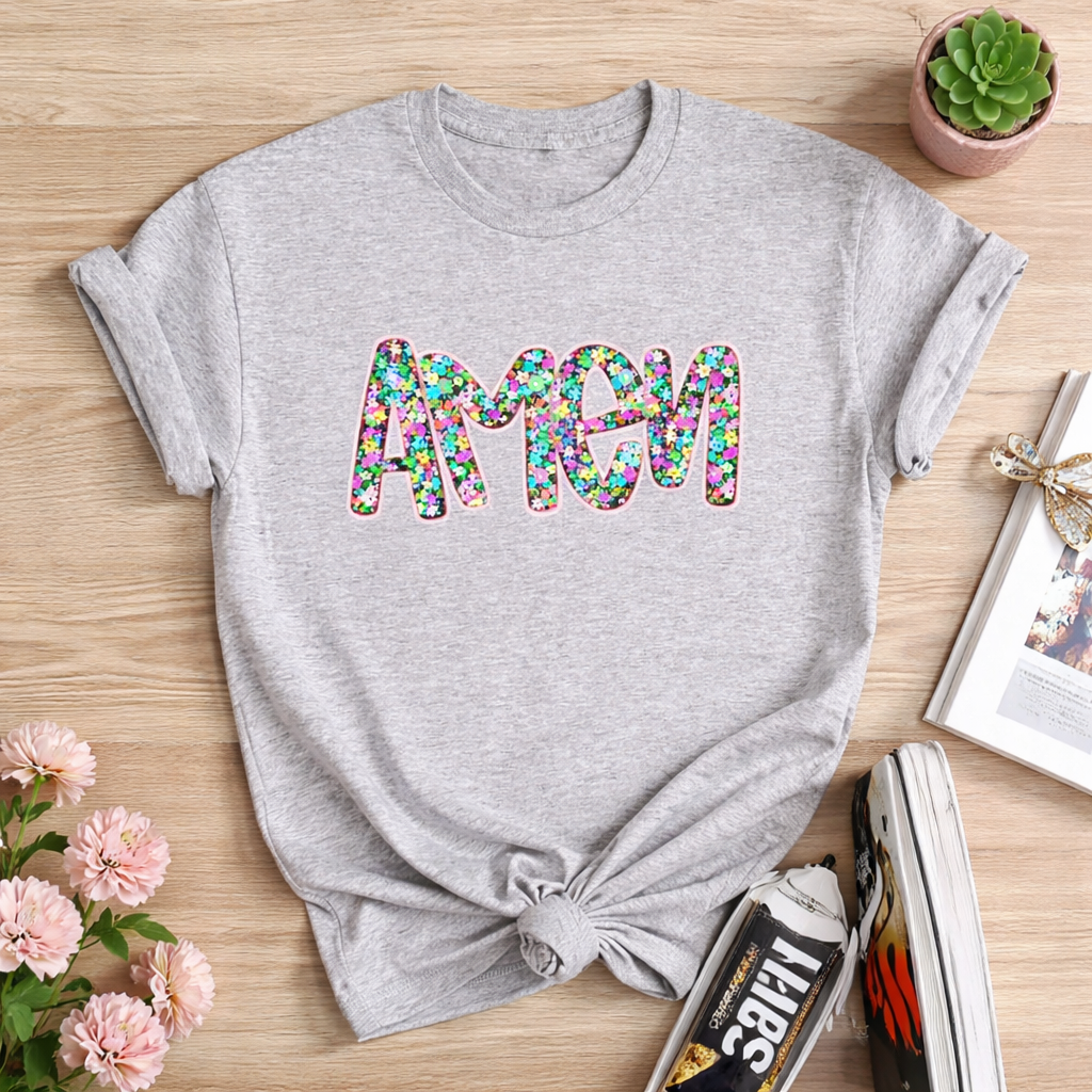 Gray t-shirt with colorful 'amen' text on a wooden surface with flowers and books.