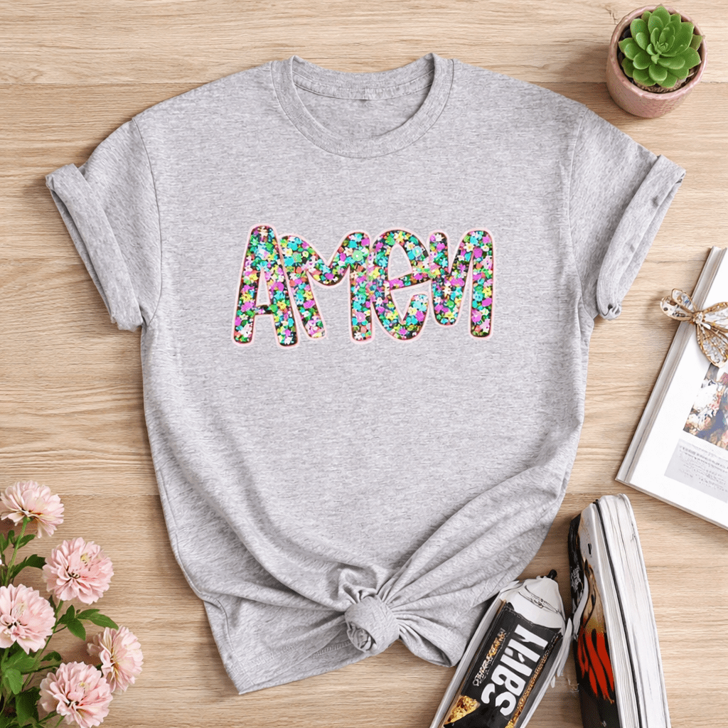 Gray t-shirt with colorful 'amen' text on a wooden surface with flowers and books.