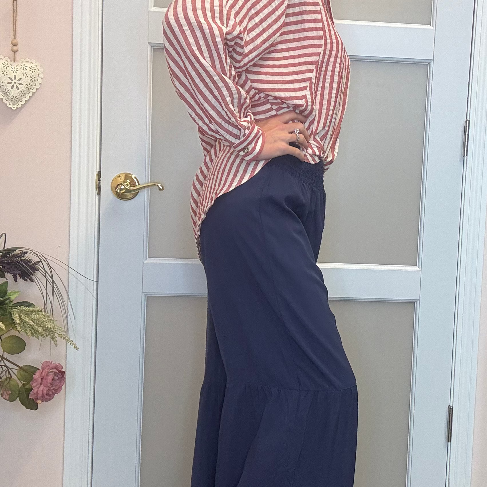 A woman posing in a pink and white striped long-sleeve top with navy blue tiered maxi pants, standing in front of a white door in a boutique setting.