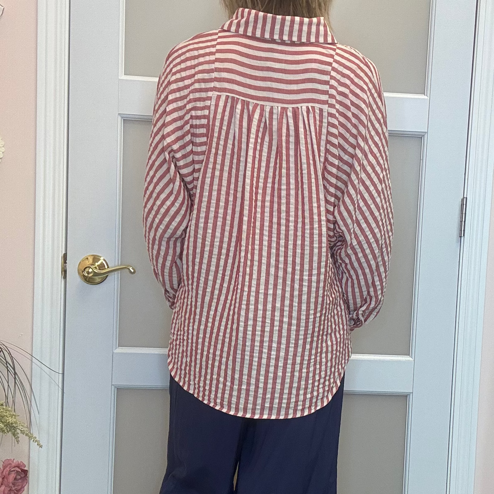 A person viewed from behind wearing a red and white vertically striped button-up shirt and navy wide-leg pants, standing in front of a white door in an indoor room.