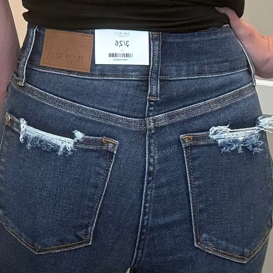 A woman viewed from behind wearing a black short-sleeve t-shirt and high-waisted blue jeans with distressed back pockets, standing indoors.