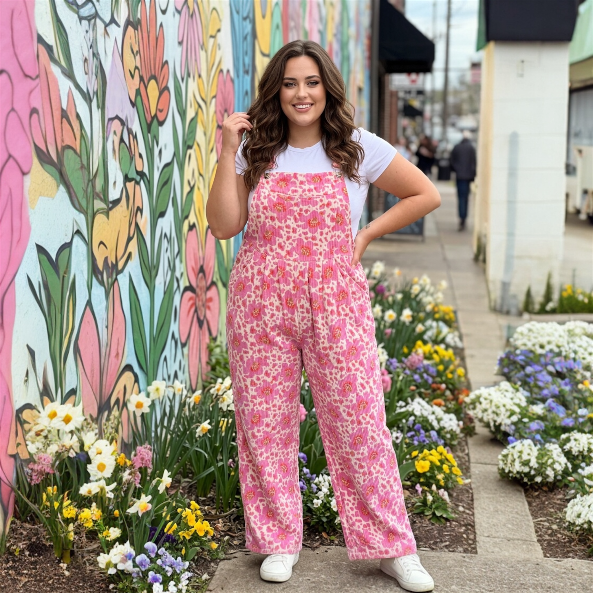 Woman in pink floral overalls standing in front of a colorful mural with flowers around.