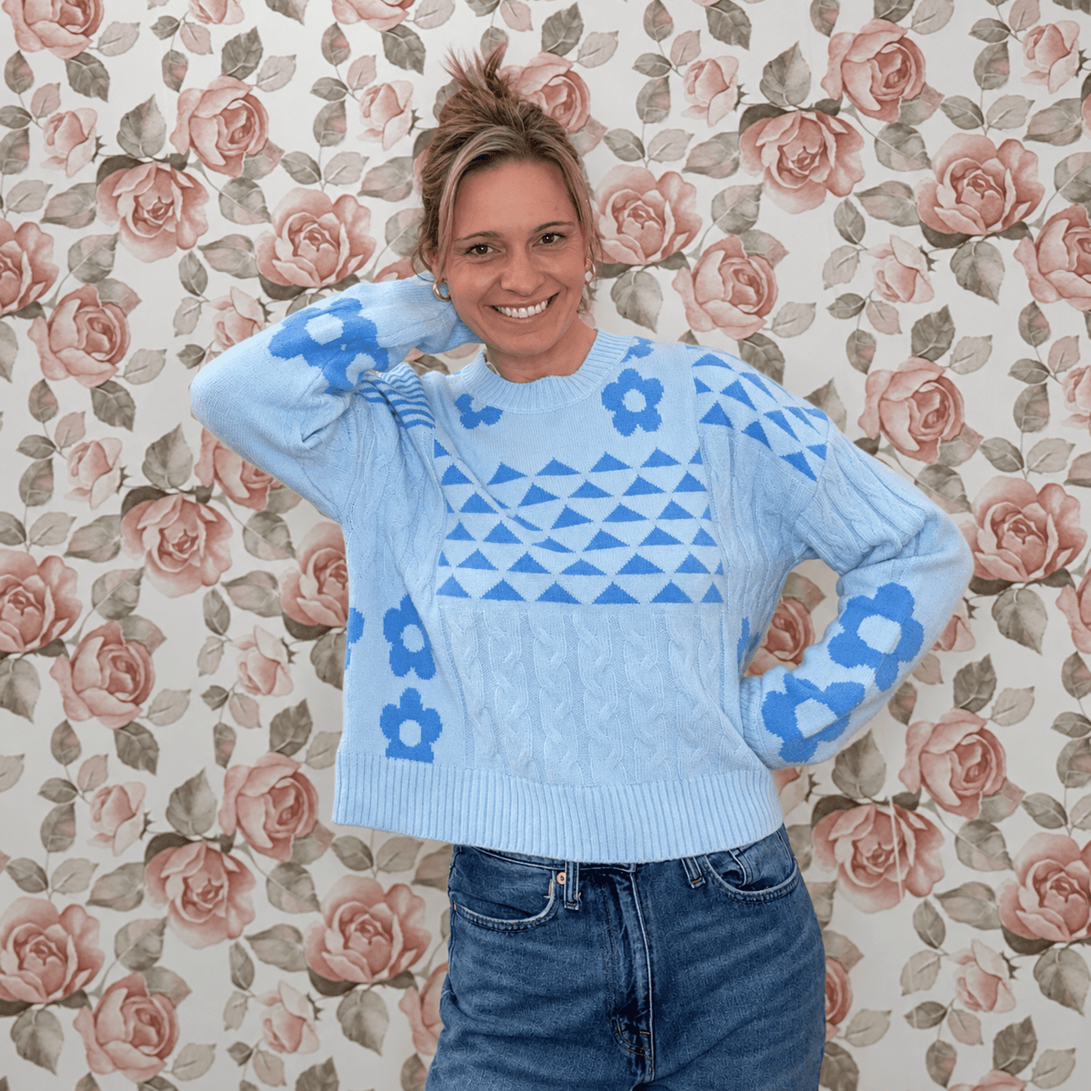 Woman wearing a light blue floral knit sweater top with blue daisy and geometric triangle pattern, paired with medium wash jeans