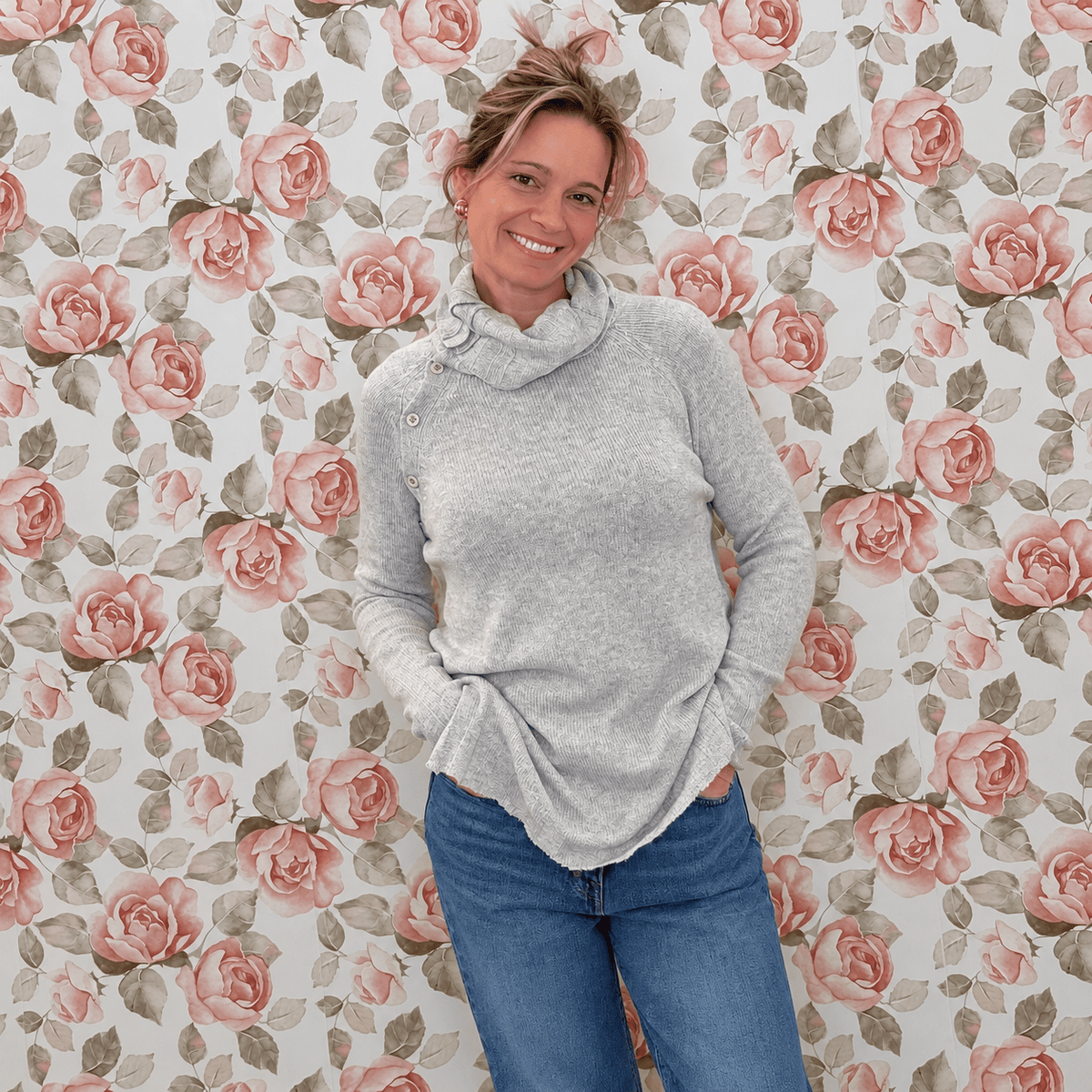Crystal wearing a heather grey cowl neck button sweater in a relaxed tunic length with jeans, in front of rose wallpaper