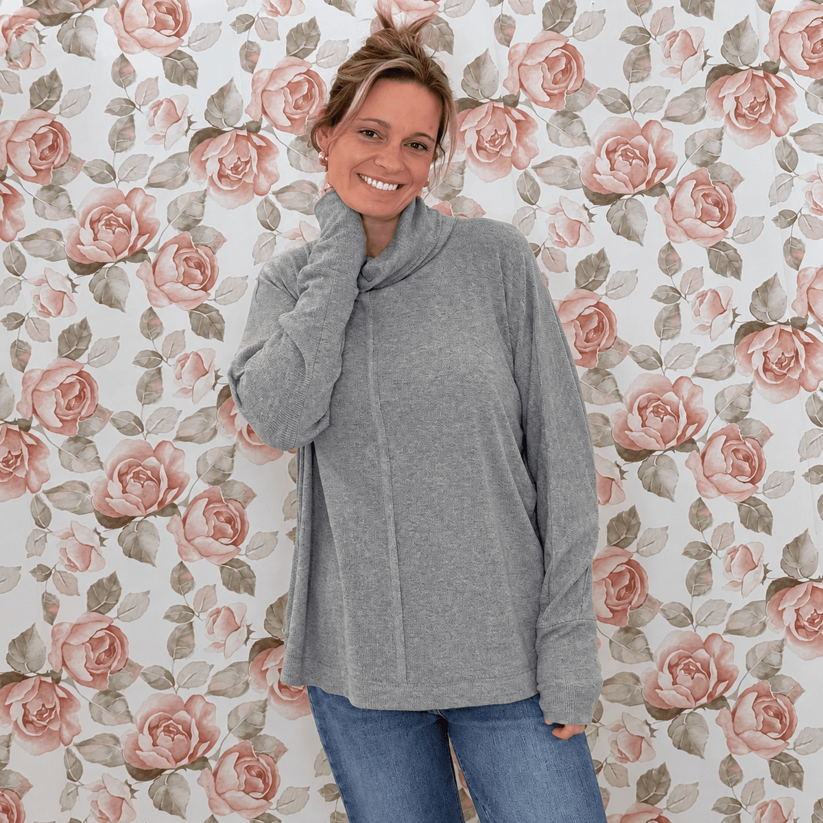 Crystal wearing a heather gray mock neck dolman tunic top with jeans, in front of rose wallpaper