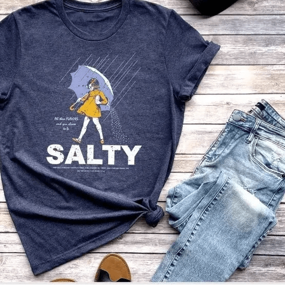 salty salt girl on a navy tshirt. shown with denim pants on.a wooden background