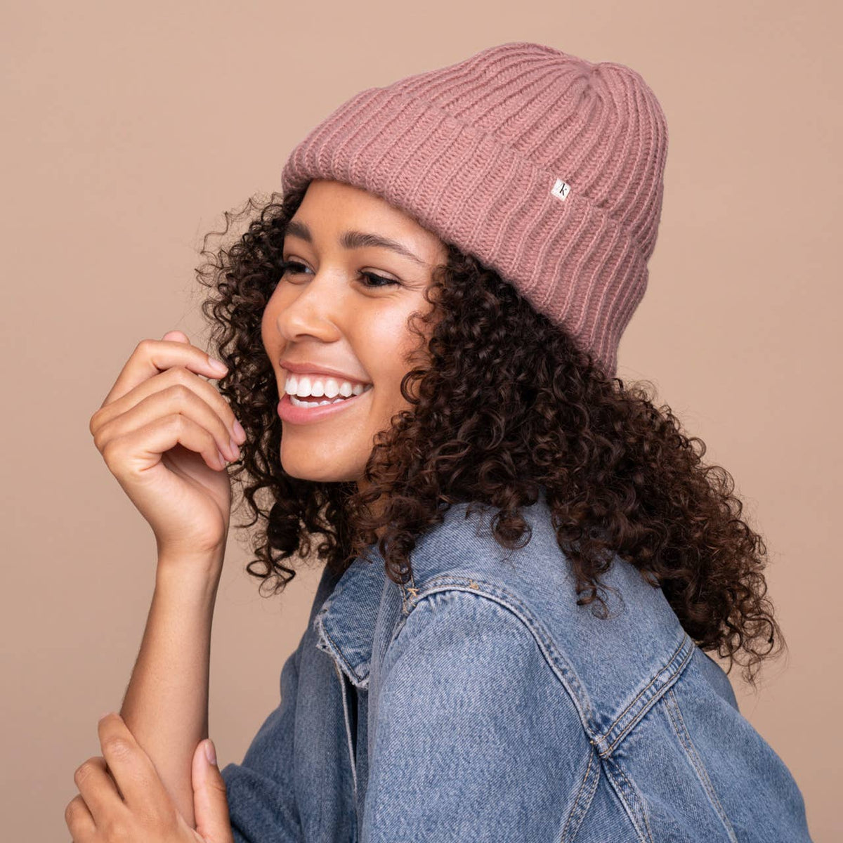 Smiling woman with curly hair wearing a blush pink KITSCH beanie and a denim jacket