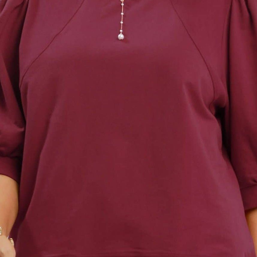 A woman wearing a burgundy crewneck sweatshirt with voluminous puff sleeves and blue jeans, shown from the chest down against a plain white background.
