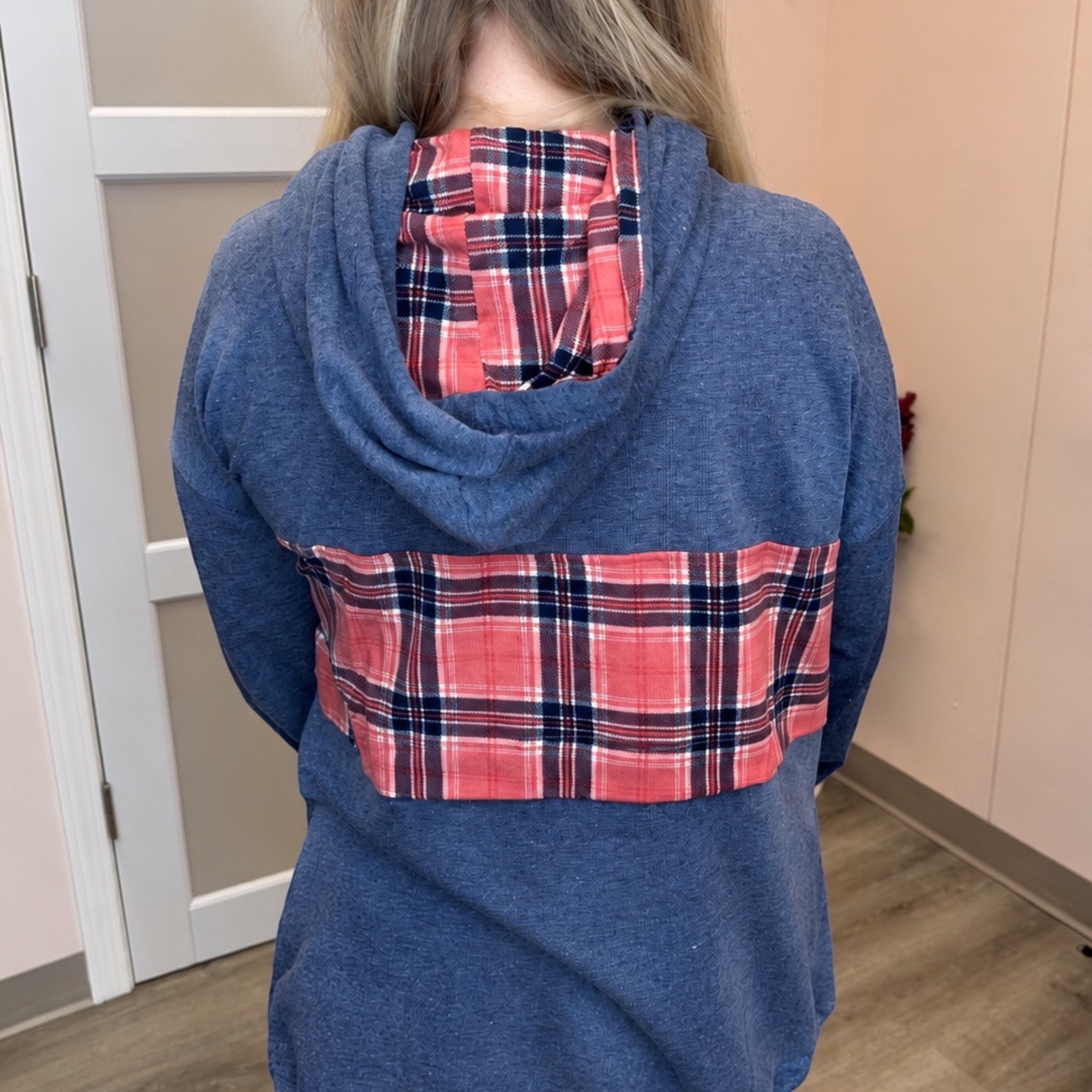 A person shown from the back wearing a heathered blue hooded sweatshirt with a red, navy, and white plaid pattern across the back and a matching plaid lining inside the hood.