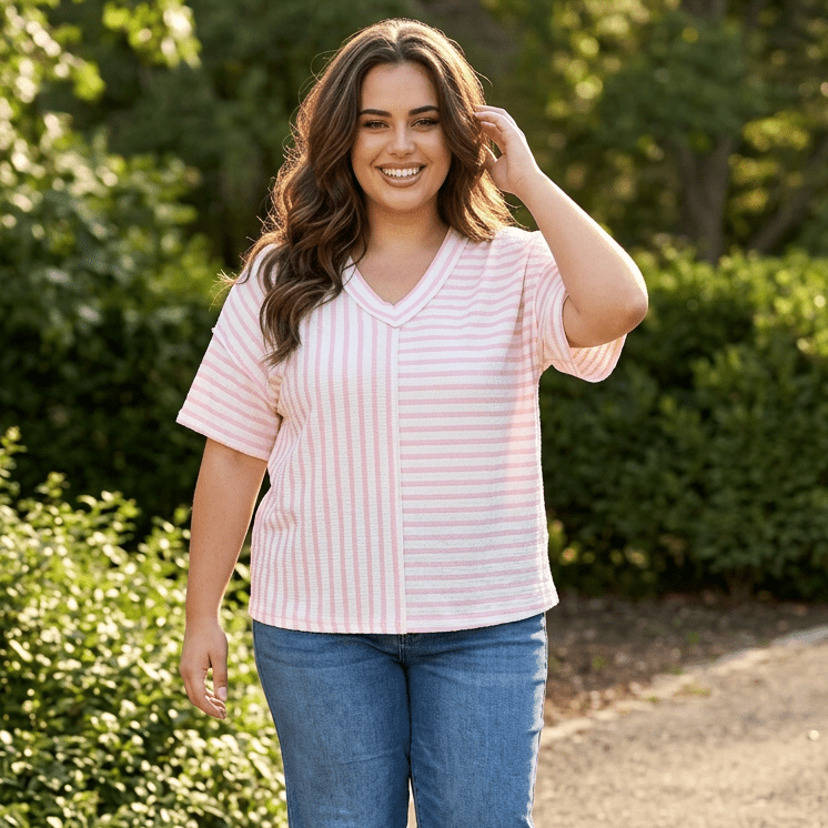 Woman wearing a pink striped shirt and blue jeans standing outdoors with greenery in the background