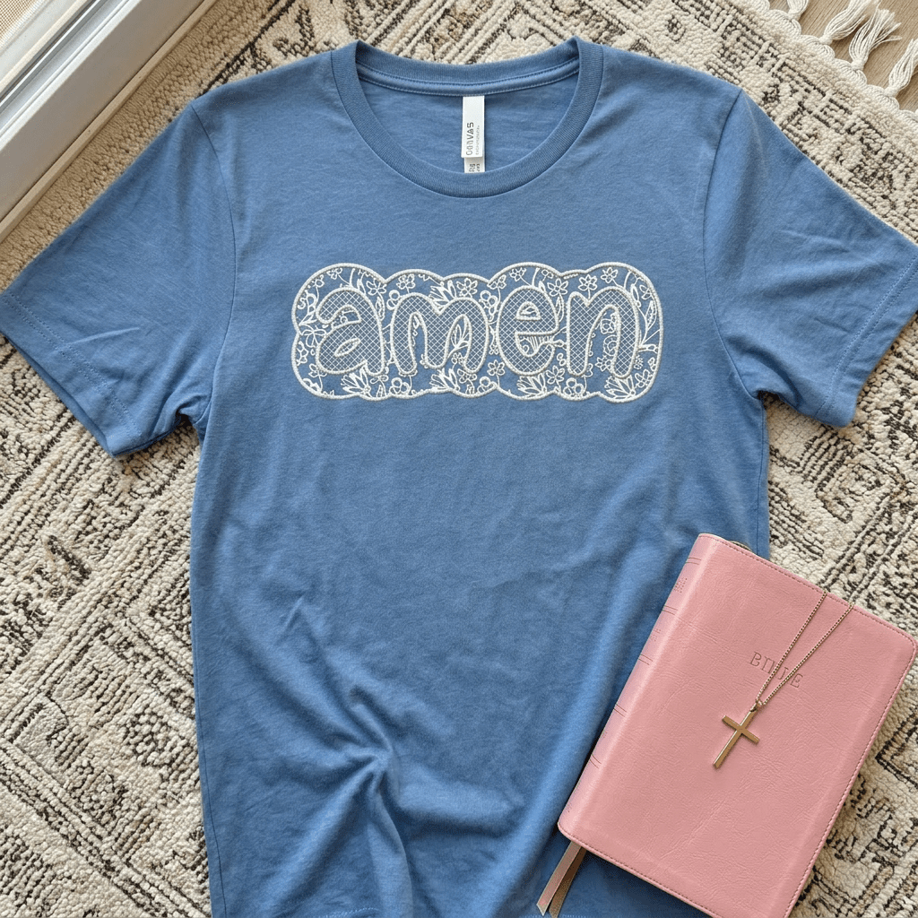 Blue t-shirt with 'amen' design on a textured surface with a pink book and cross necklace.
