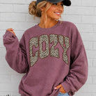 A woman wearing a heather maroon sweatshirt with a leopard-print 'COZY' graphic on the front, paired with a black baseball cap and light-wash jeans, standing against a white brick wall. Text in the bottom-left corner reads 'Heather Maroon'.