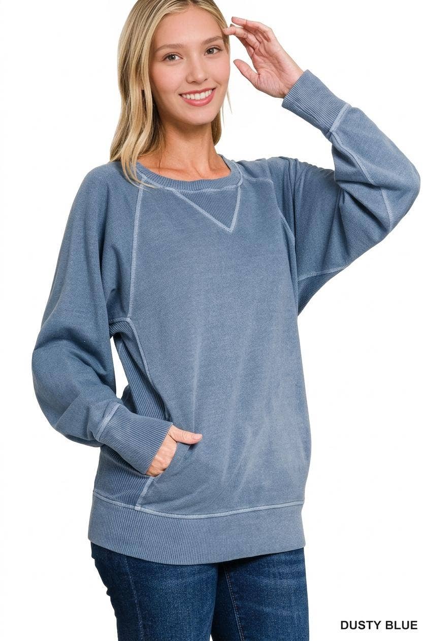 Woman wearing a washed dusty blue French terry pullover with raglan sleeves, V-stitch neckline, contrast stitching and front pockets, styled with dark jeans