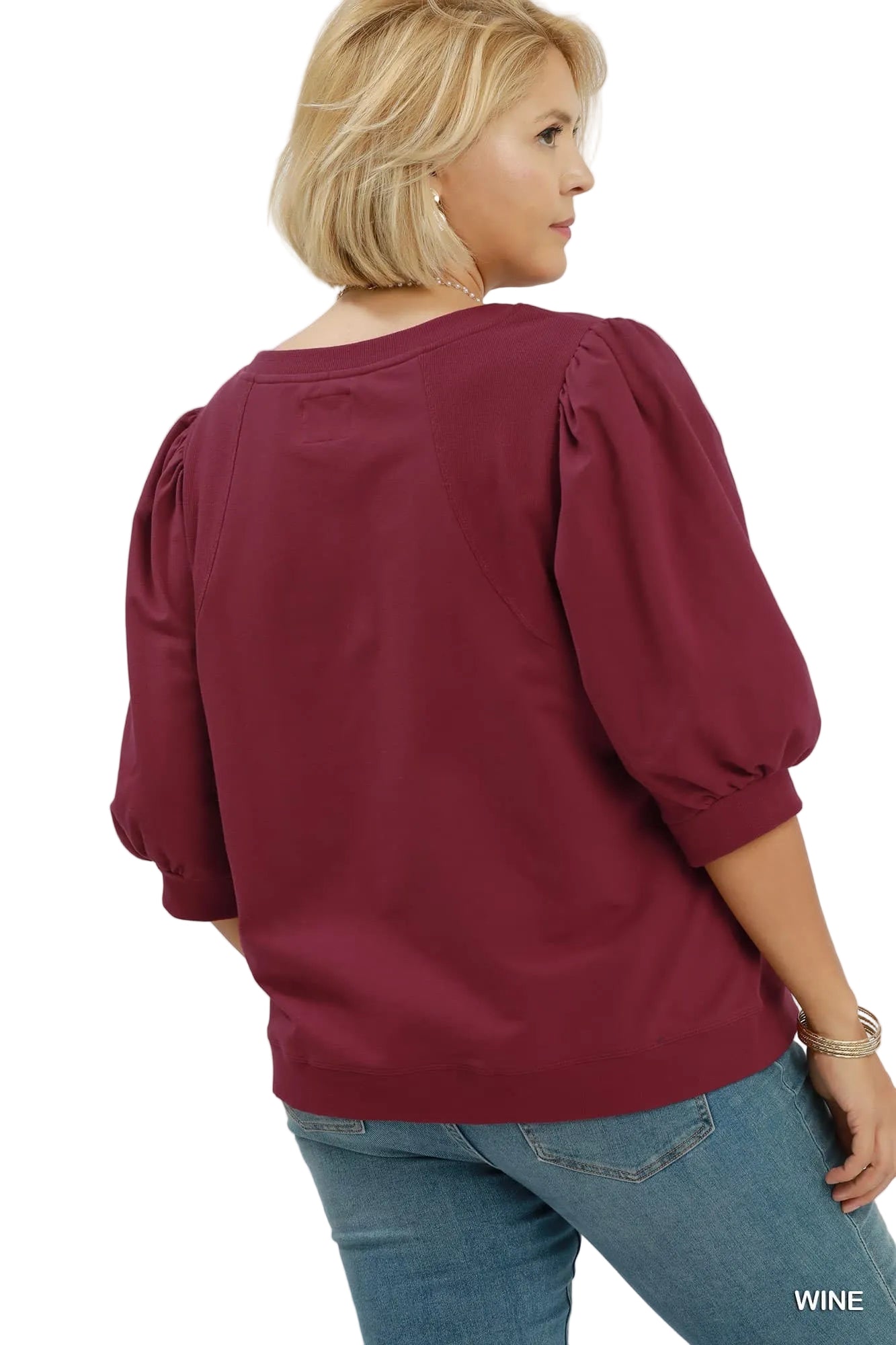 A woman shown from the back wearing a wine-colored puff-sleeve blouse and blue jeans against a light gray studio background. The text 'PLUSSIZE' appears in the top right, and 'WINE' appears in the bottom right.