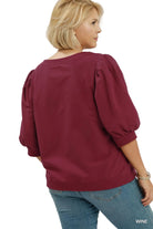 A woman shown from the back wearing a wine-colored puff-sleeve blouse and blue jeans against a light gray studio background. The text 'PLUSSIZE' appears in the top right, and 'WINE' appears in the bottom right.