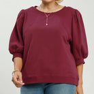 A woman modeling a wine-colored long-sleeve top with voluminous puff sleeves and a round neckline, paired with blue jeans. The image has a white background with 'PLUS SIZE' text in the top right and 'WINE' text in the bottom right.