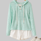 Mint green hoodie with lace-up front on a hanger, white shorts, and white sneakers on a light wooden floor.