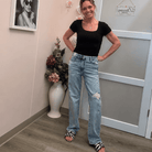 A woman posing in a black scoop-neck t-shirt and light-wash distressed jeans inside a room with pink walls, framed art, and a white door.