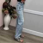 A woman stands in a room with pink walls, a white door, and a vase of flowers, wearing a black short-sleeve top and light-wash wide-leg jeans.