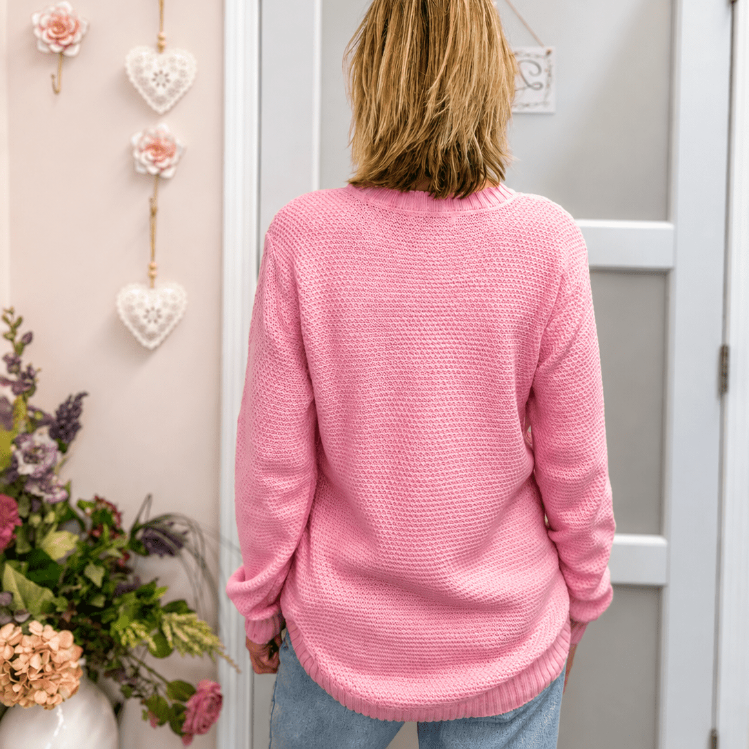 A woman viewed from behind wearing a pink textured knit sweater and light blue jeans, standing in front of a white door in a decorated room with pink walls and floral accents.