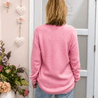 A woman viewed from behind wearing a pink textured knit sweater and light blue jeans, standing in front of a white door in a decorated room with pink walls and floral accents.