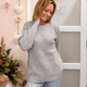 A woman wearing a light gray waffle-knit sweater and light blue jeans, standing in front of a white door with a pink wall decorated with hanging flowers and hearts.