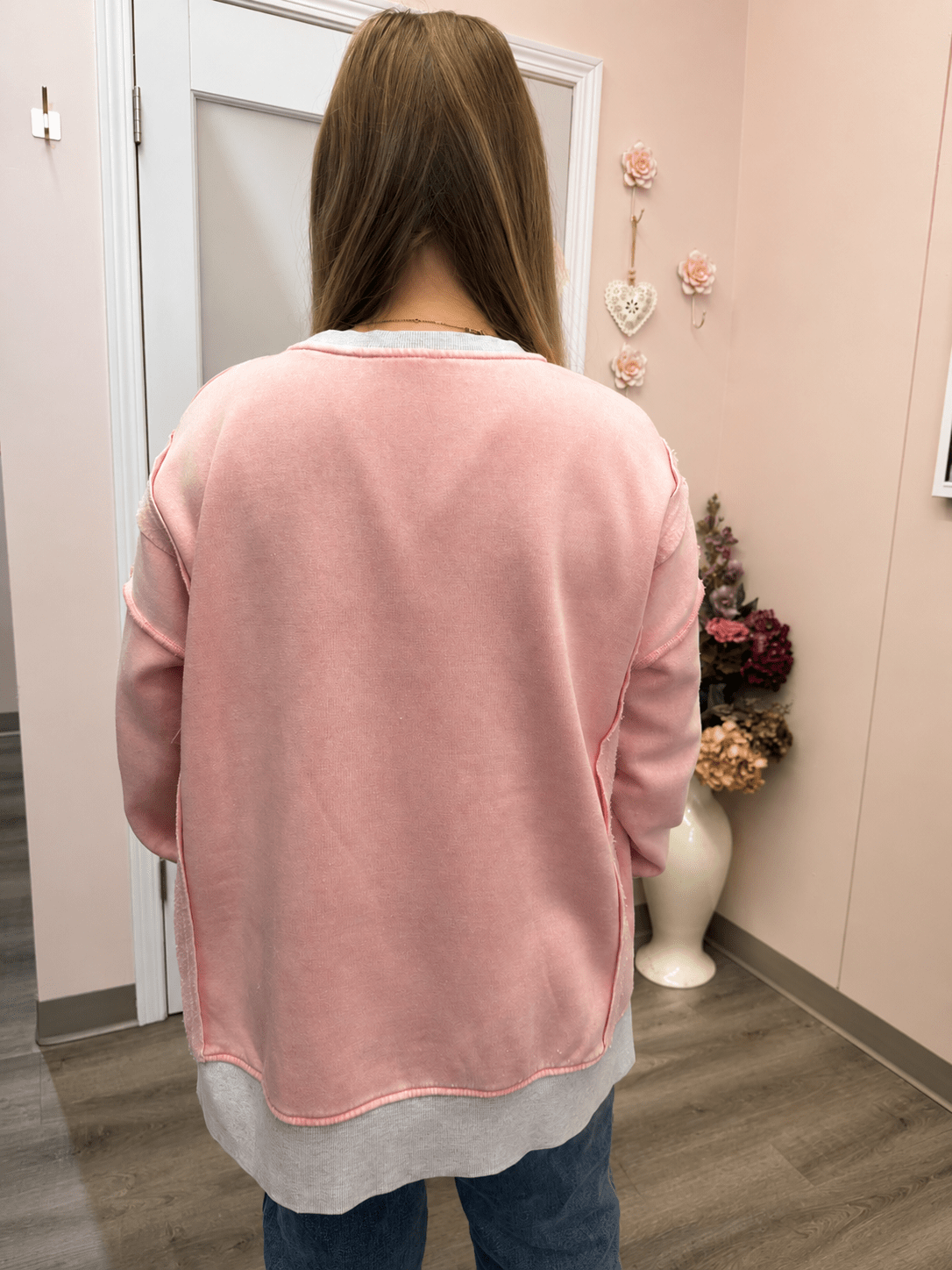 A woman viewed from behind wearing a pink crewneck sweatshirt with a light gray undershirt and blue jeans, standing in a room with pink walls and a white door.