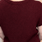 A woman viewed from behind wearing a short-sleeve ribbed knit sweater in burgundy and blue jeans, standing in a room with a pink wall, wooden floor, and a vase of flowers.