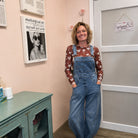A woman stands indoors wearing a brown floral long-sleeve shirt under blue denim overalls. The room has peach walls, framed wall art, a white door, and a teal cabinet.
