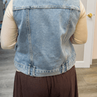 A person viewed from behind wearing a light blue denim vest over a beige long-sleeve top and dark brown pants, standing indoors near a white door.